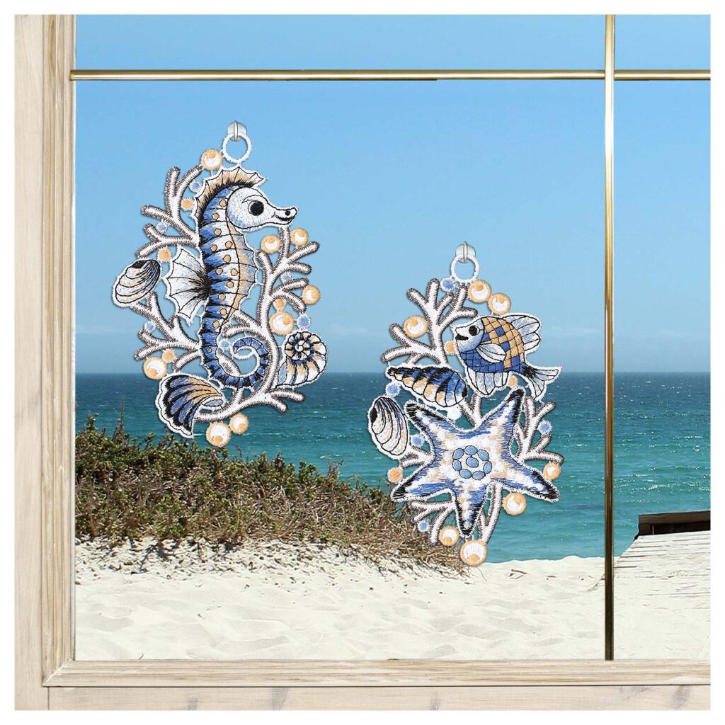 Zwei gestickte Sonnenfänger - ein blaues Seepferdchen und ein blauer Fisch mit einem Seestern - hängen an einem Fenster mit Blick auf einen Sandstrand, grüne Dünen und das Meer unter einem klaren Himmel.