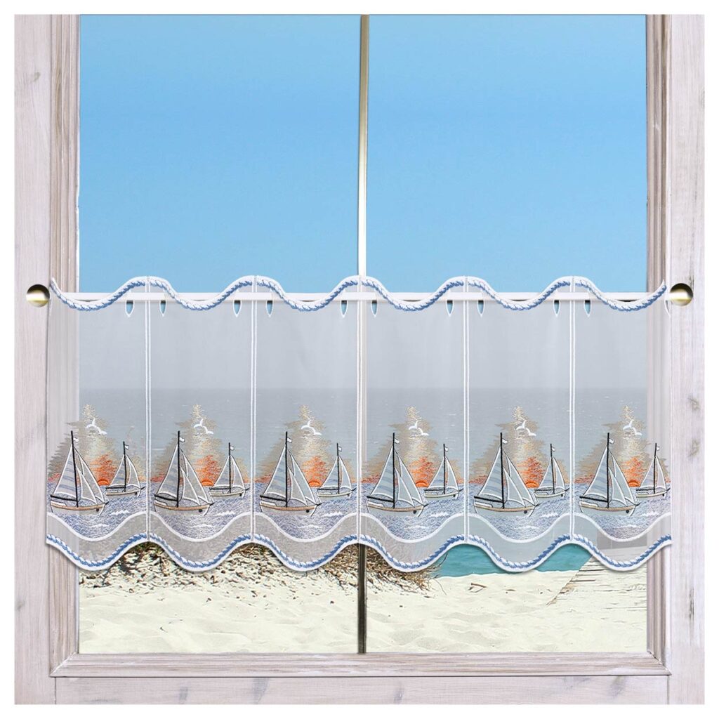 Ein Fenster mit durchsichtigen, bestickten Vorhängen, auf denen Segelboote und Möwen abgebildet sind. Draußen befindet sich ein Sandstrand, im Hintergrund sind das blaue Meer und der klare Himmel zu sehen.