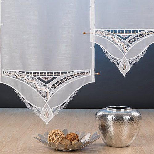 Vor einer dunklen Wand hängen zwei weiße, spitzenbesetzte Vorhänge mit verschlungenen Mustern. Auf dem hellen Holzboden darunter ruht eine Metallvase neben einer dekorativen Schale mit geflochtenen Bällen - eine inspirierende Szene für jede moderne Webagentur oder jedes E-Commerce-Studio.