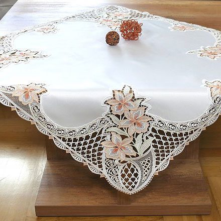 Ein Holztisch ist mit einer dekorativen weißen Tischdecke mit aufwändiger Spitzen- und Blumenstickerei bedeckt - eine einladende Szene, die oft auf E-Commerce-Websites oder von einer kreativen Werbeagentur präsentiert wird, wobei zwei kugelförmige Objekte als einfache Muster-Dekostücke dienen.