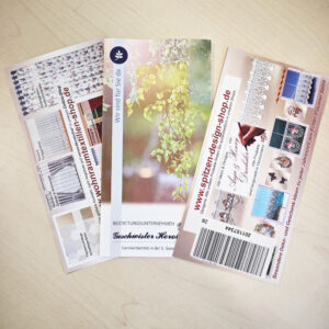 Auf einer hellen Holzfläche sind drei farbenfrohe Flyer ausgebreitet, die Bilder von Gardinen, Spitzenmustern und einem Baum mit Sonnenlicht zeigen, jeweils mit deutschem Text und Angaben zur Website.