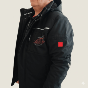 Ein Mann, der eine schwarze Kapuzenjacke mit roter Stickerei und einem kleinen roten quadratischen Aufnäher am Ärmel trägt, steht vor einem schlichten hellen Hintergrund.