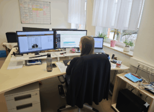 Eine Frau arbeitet an einem Schreibtisch mit drei Computermonitoren, Papierkram und Büromaterial. Große Fenster mit Jalousien lassen natürliches Licht herein, und mehrere Topfpflanzen stehen auf der Fensterbank. In der Nähe hängt ein Wandkalender.