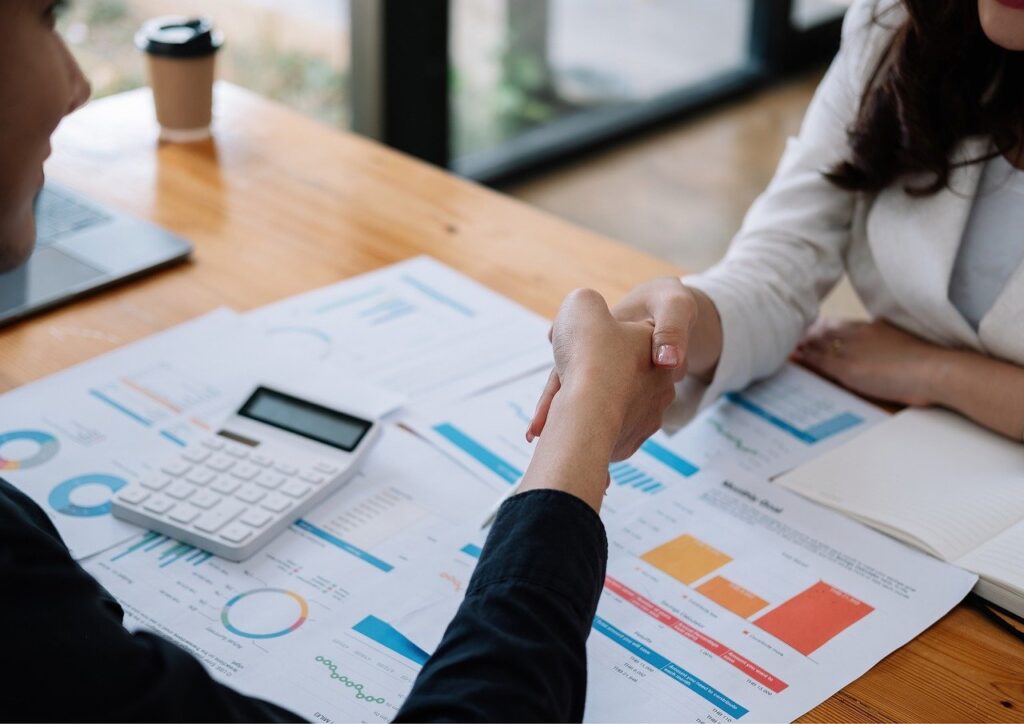 Zwei Personen schütteln sich die Hände über einem Schreibtisch, der mit Finanztabellen, Diagrammen, Dokumenten, einem Taschenrechner und einem Notizbuch bedeckt ist, und unterzeichnen damit eine Geschäftsvereinbarung über Fördermittel oder Zuschüsse für sächsische KMU. Im Hintergrund steht eine Kaffeetasse.